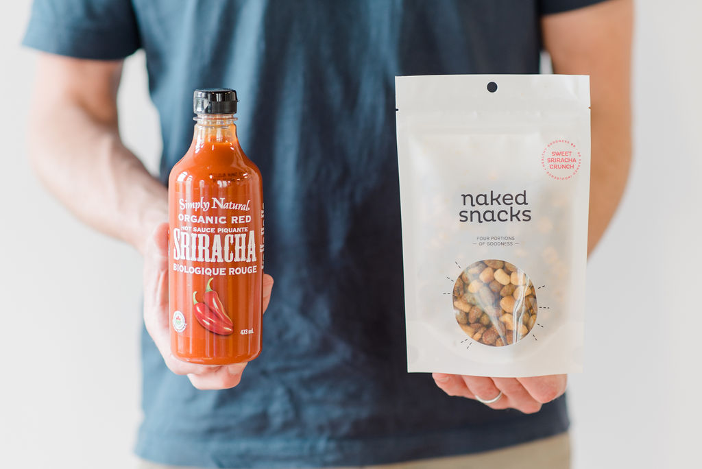 Man holding a bag of Naked Snacks Sweet Sriracha Crunch and a bottle of Simply Natural Organic Red Sriracha Flavour