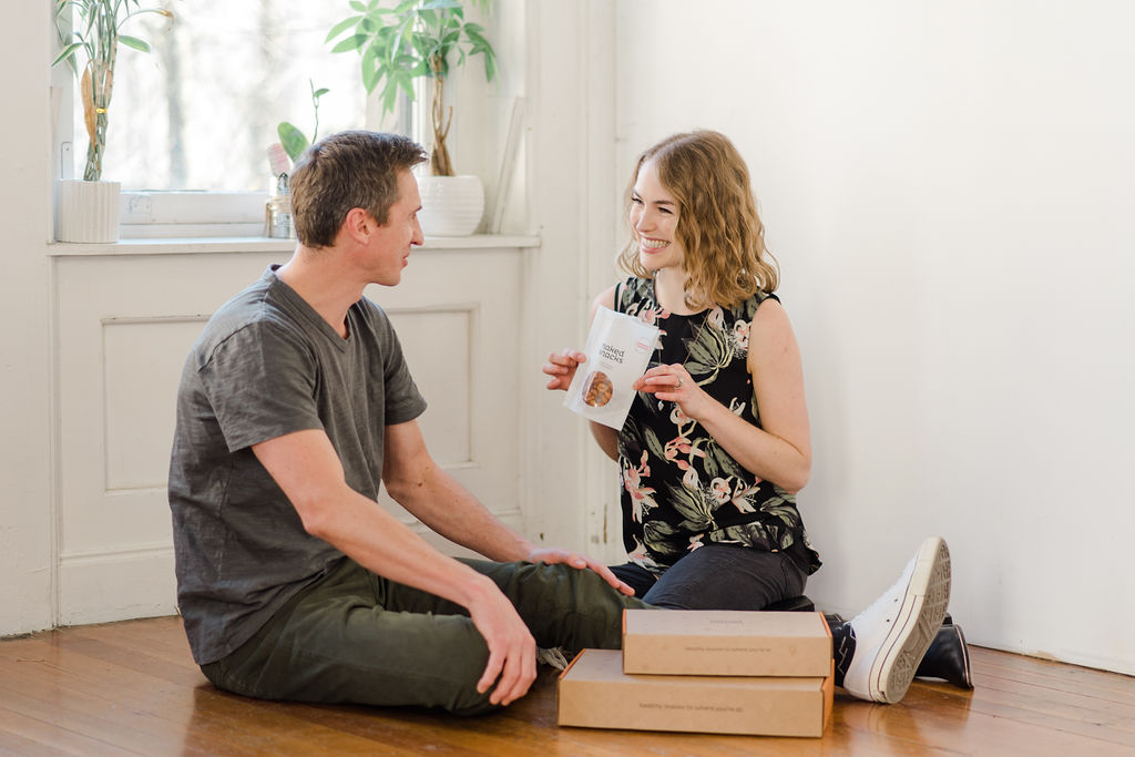 Neil Thomson and Ryley Humphry, co-owners of Laid Back Snacks, sitting on the floor with 2 boxes of Naked Snacks