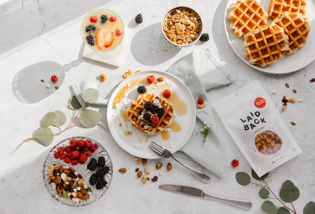 Laid Back Snacks Hearty Tamari laying on white table with berry waffles, a tray of snacks and berries and mimosa