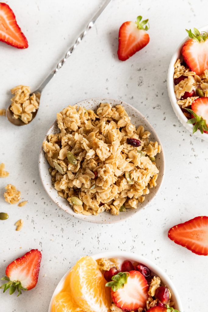 granola in a white bowl on a white countertop with fruits, strawberries and oranges surrounding it